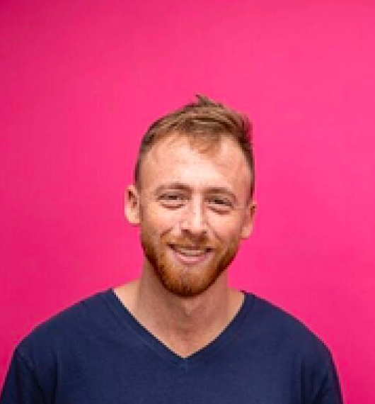 Smiling man with light brown hair and beard wearing a navy blue v-neck shirt against a pink background.