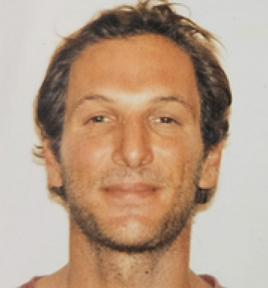 Close-up of a smiling man with short brown hair and light facial stubble against a plain background.