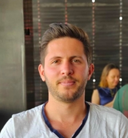 Young man with short brown hair and light beard wearing a white shirt seated indoors with a blurred background showing another person.