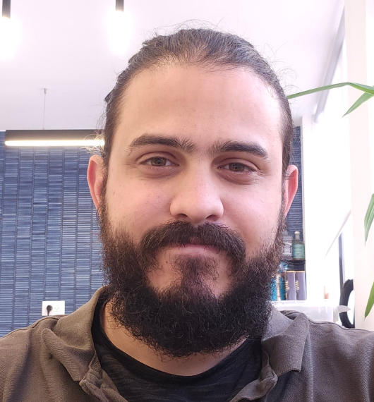Man with dark hair tied back and a full beard smiling slightly in an indoor setting with a blue tiled wall and some plants in the background.