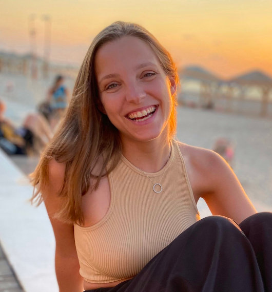 Young woman smiling with long hair, wearing a beige sleeveless top and a circular pendant necklace at sunset on a beach.