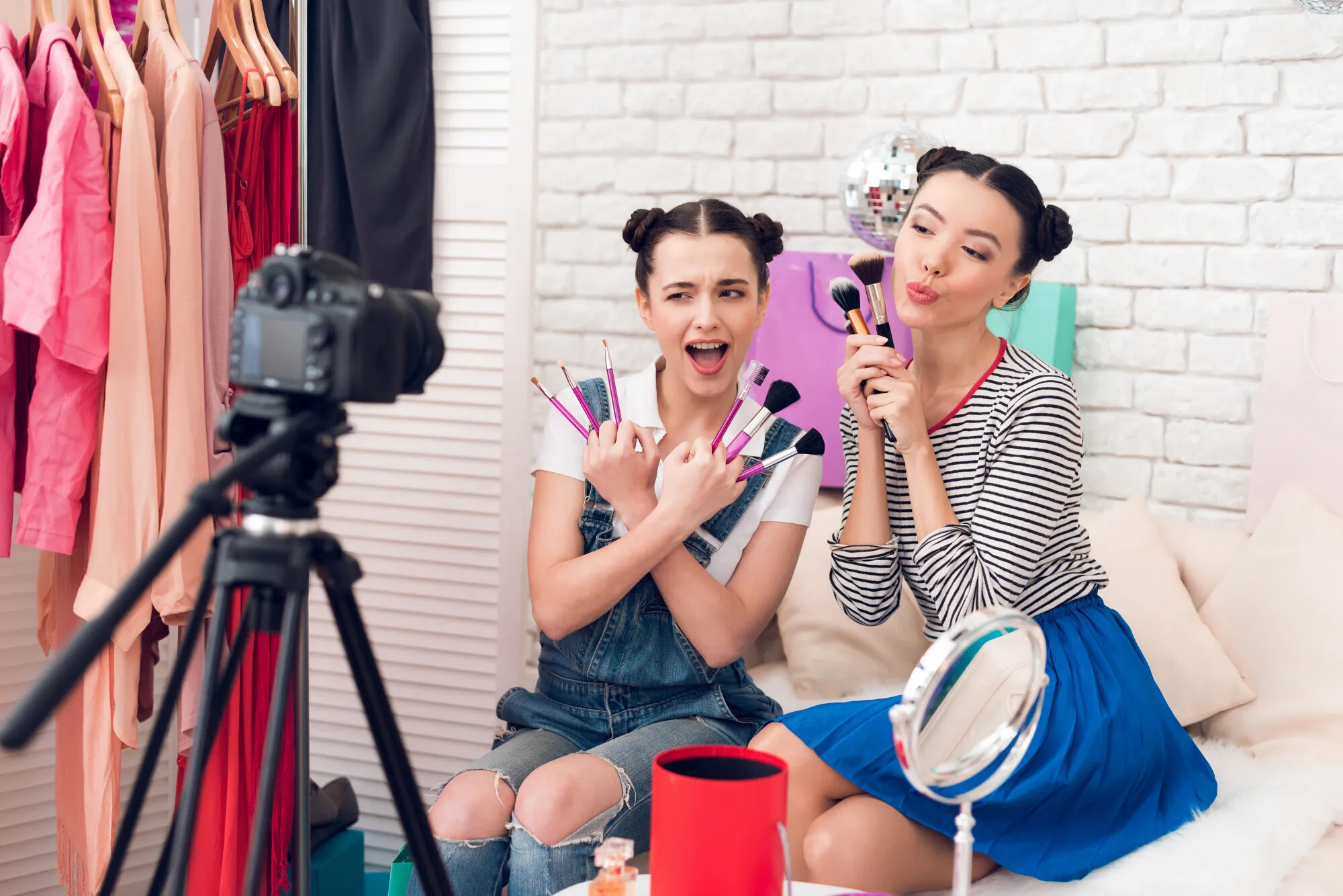 Two young women with hair buns holding makeup brushes posing for a camera in a bedroom with clothing rack and mirror.