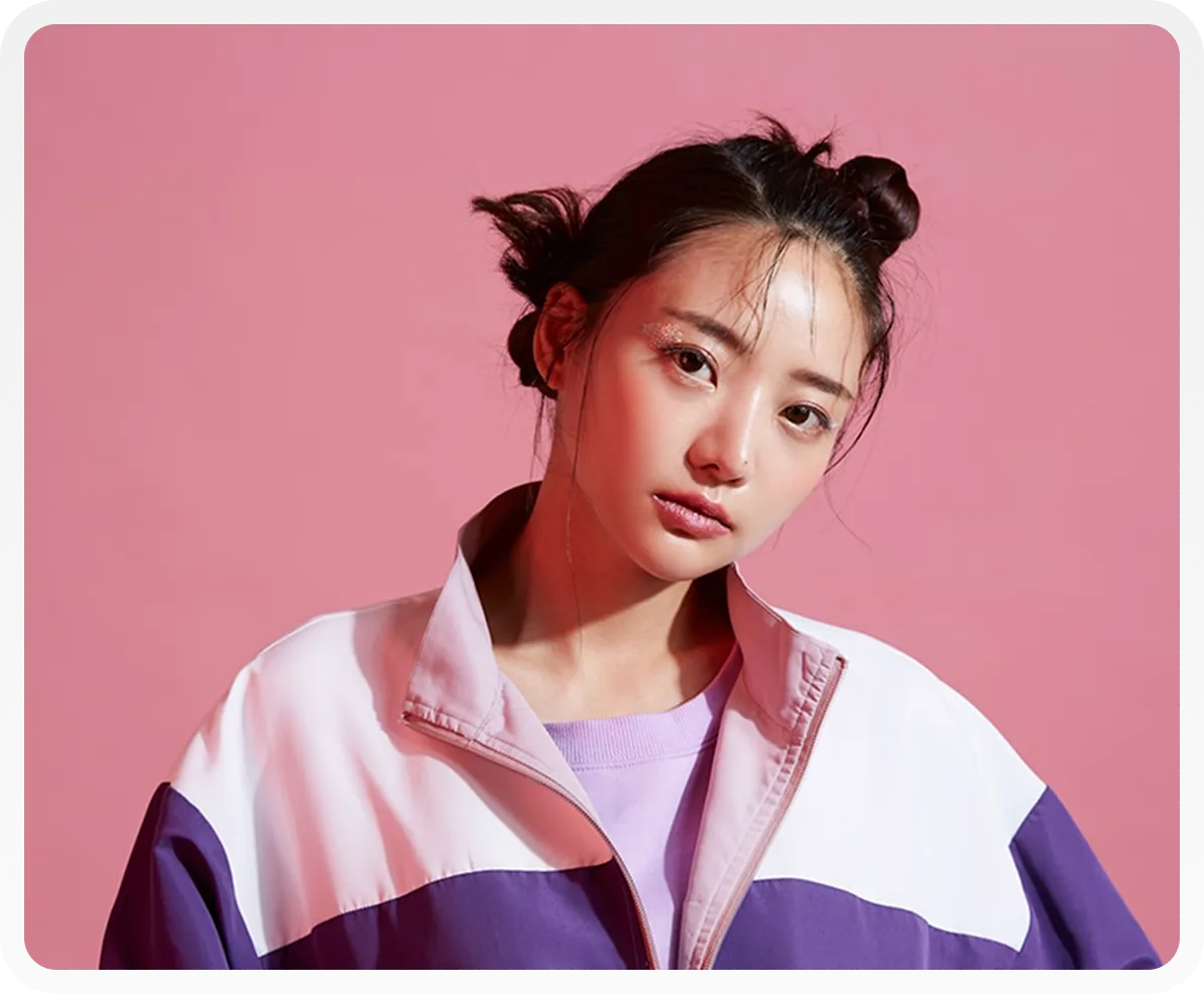 Young woman with two buns and glitter eye makeup wearing a purple and white jacket against a pink background.