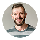 Smiling man with short brown hair and beard wearing a gray striped shirt against a light background.