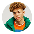 Young man with curly brown hair wearing a green jacket and orange sweater, looking directly at the camera against a plain background.