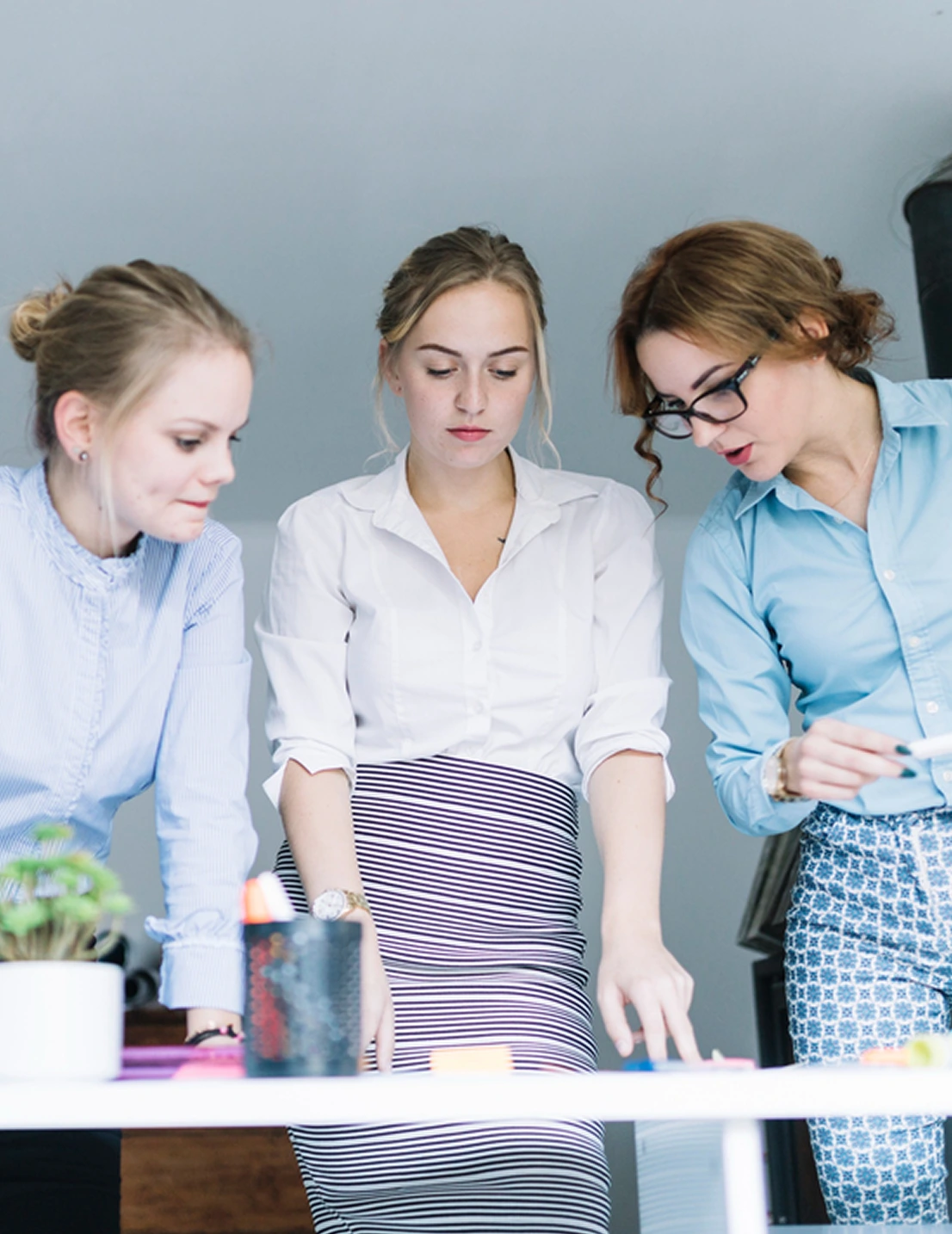 Three women engaged in discussion and pointing at items on a table in a bright office.