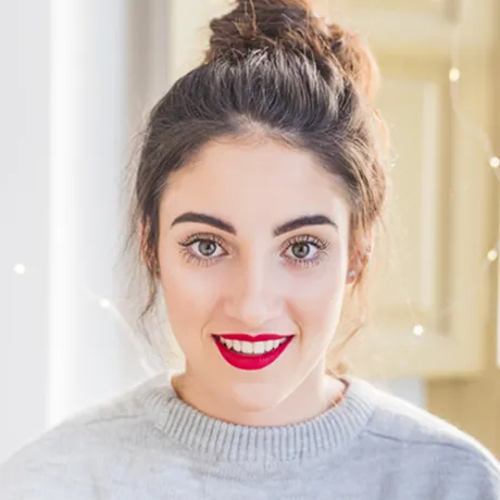 Young woman with dark hair in a bun, wearing red lipstick and a light gray sweater, smiling indoors.