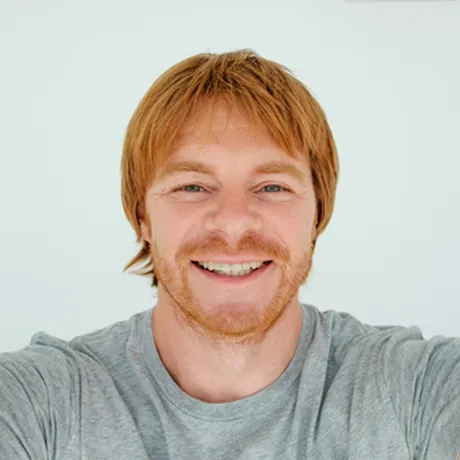 Smiling young man with red hair and beard wearing a gray shirt against a plain light background.