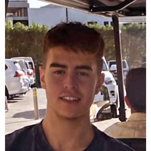 Young man with short brown hair smiling, sitting outdoors with parked cars in the background.