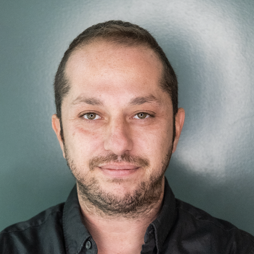 Headshot of a man with short hair and light facial hair wearing a dark shirt against a teal background.